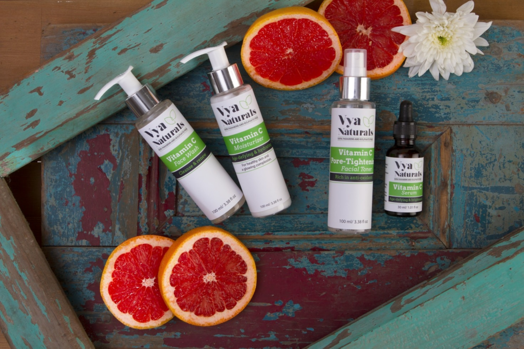 Linha de skincare vegano Vya Naturals com Vitamina C — sabonete facial, hidratante, tônico e sérum sobre bandeja de madeira com fatias de toranja